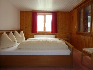 A wooden-paneled bedroom with a bed, pillows, a window with red curtains, and a wooden chair. - Cozy Vacation Rental at the Schwendehof Vacation Home (Fontanella)