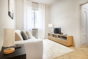 Ein modernes Wohnzimmer mit einer Couch, einem Fernseher auf einem Holzschrank, einer Lampe und einem Fenster mit Vorhängen.