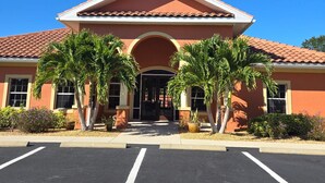 A building with a red facade, a tiled roof, and a central entrance with a semi-circular arch. There are palm trees in front and a parking lot with marked spaces. - Cozy 2-bedroom condo in nice Fort Myers (Fort Myers)