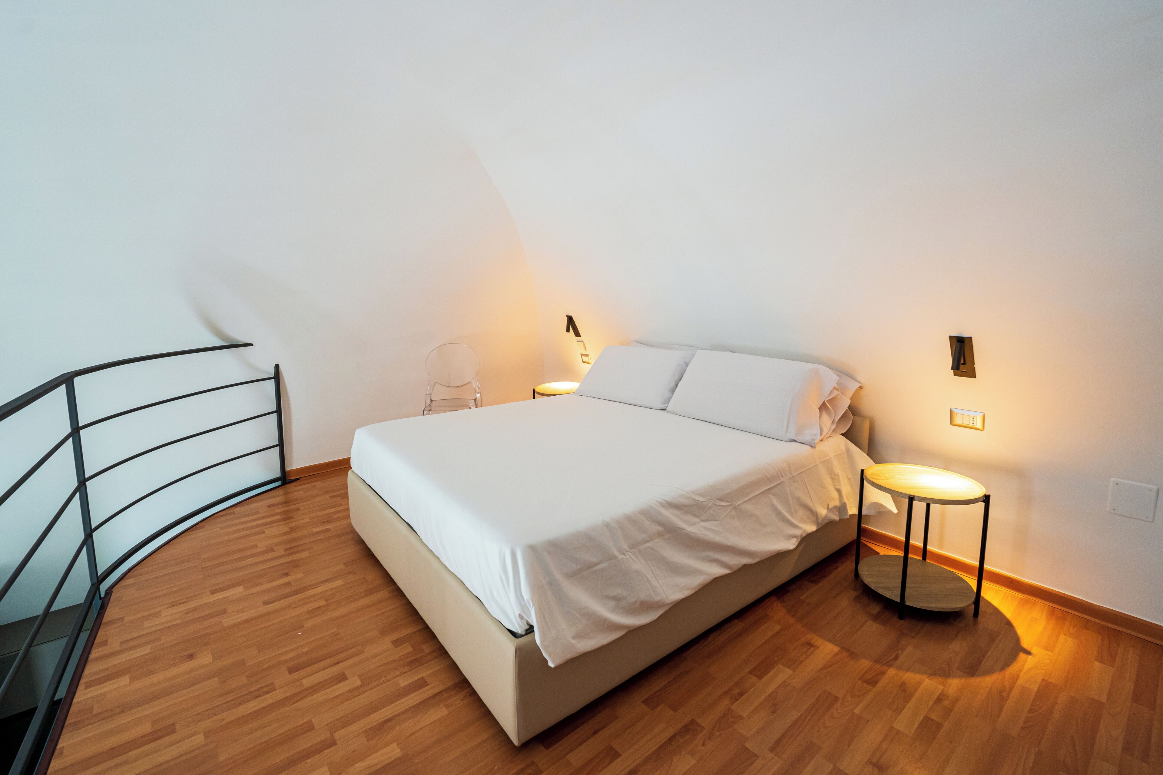 A bedroom with a bed, a bedside table, a chair, and a small table with a lamp.
