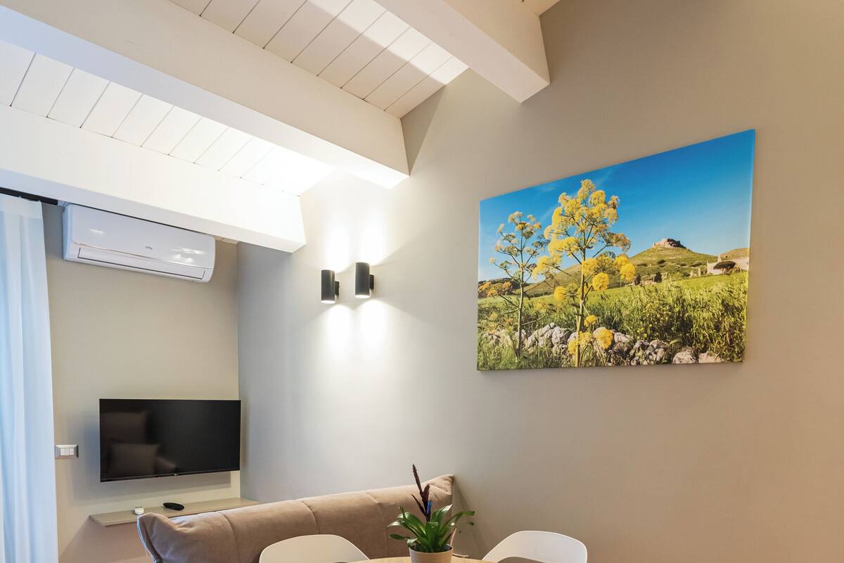 A modern living room with a round wooden table, white chairs, a beige sofa, a flat-screen TV, and a painting of a landscape.