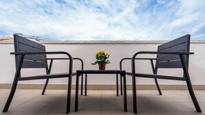 A rooftop terrace with two black metal chairs and a small table, a potted plant with yellow flowers, and a clear sky.