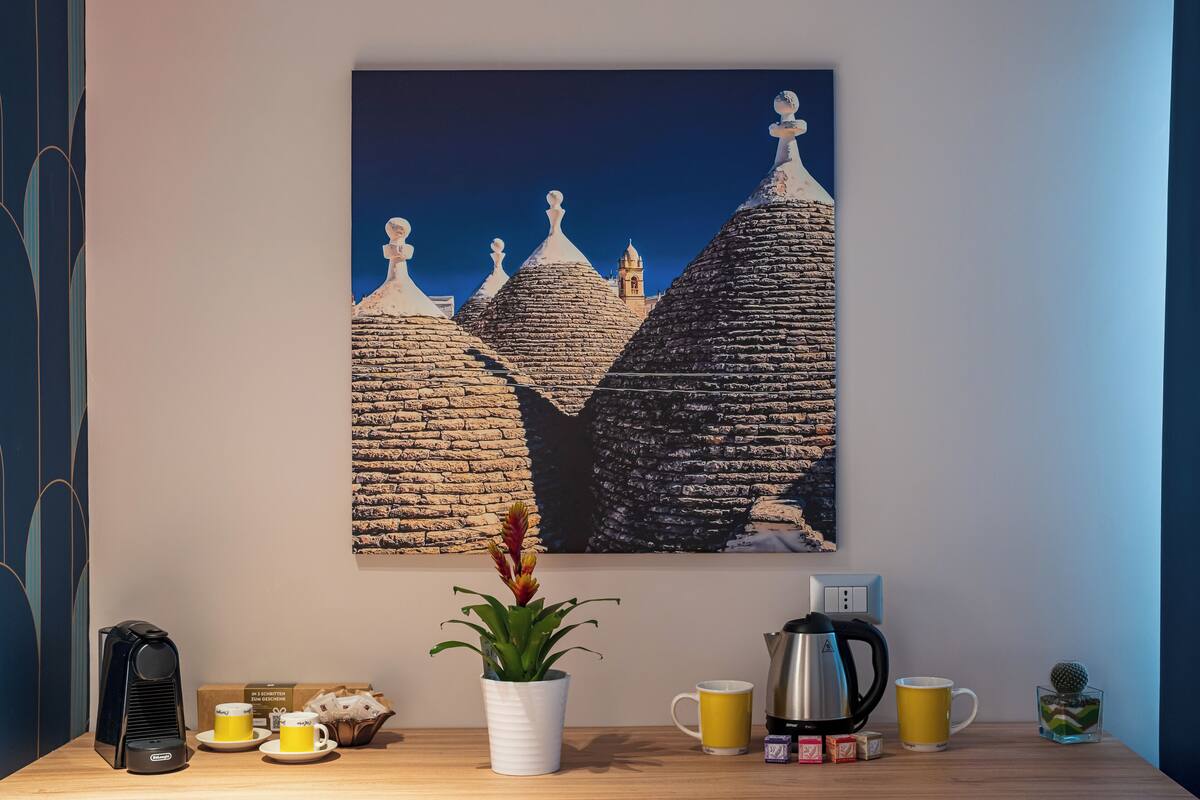 A wall-mounted painting of traditional architecture, a coffee maker, a potted plant, and a mug on a wooden table.