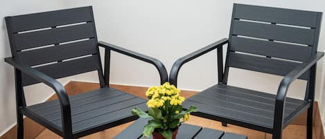 Un balcón con dos sillas de metal negro y una mesita, una planta en maceta con flores amarillas, y un edificio blanco al fondo.
