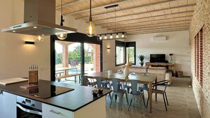 A modern kitchen with a central island, dining area with chairs, a TV, and a stone wall. - The Private Accommodation Villa in Peralada (Peralada)