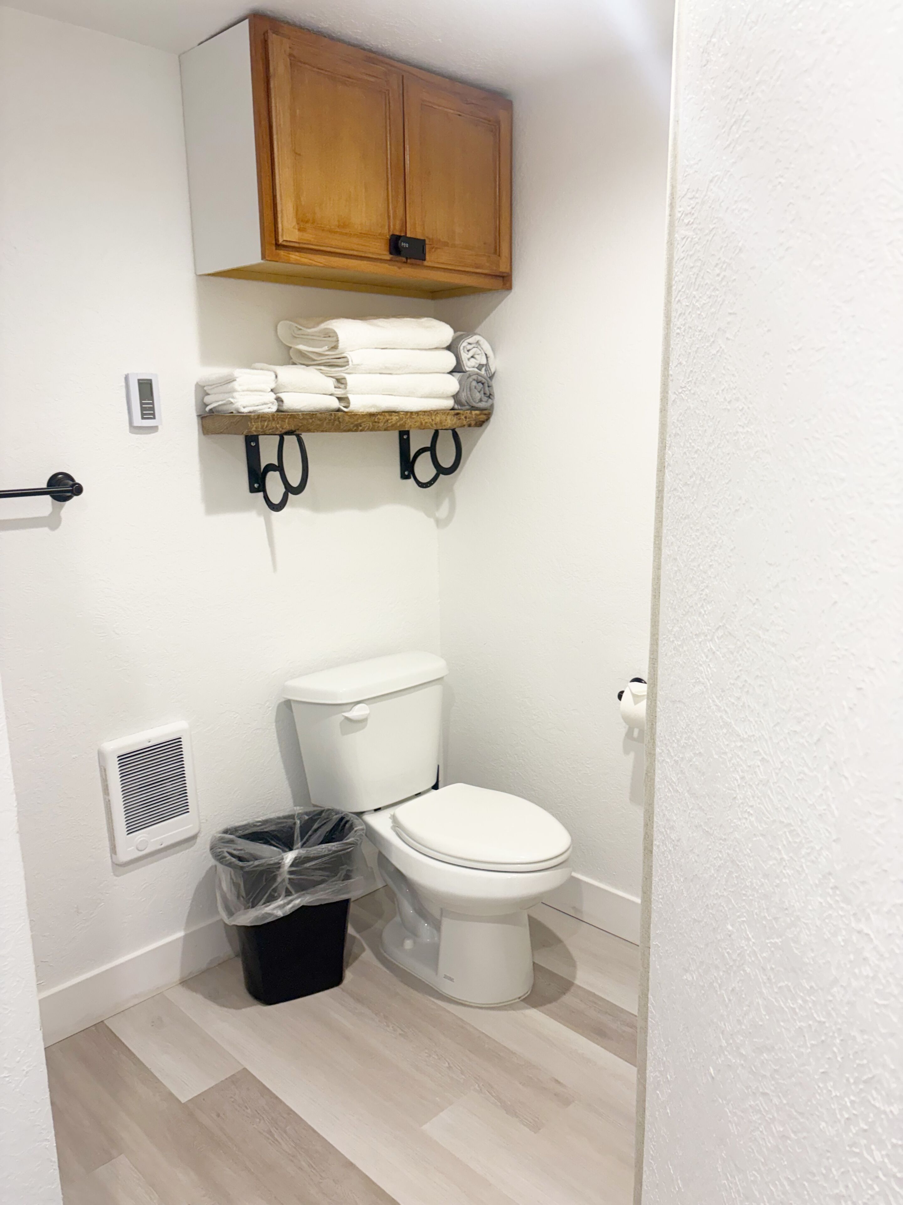 Ein kleines Badezimmer mit Toilette, einem Holzschrank über dem Waschbecken, einem Handtuchhalter mit Handtüchern, einem Mülleimer und einem Lüftungsgitter.