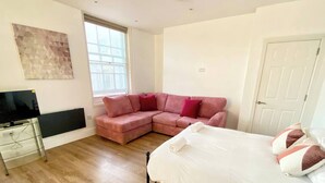 A bedroom with a bed, a pink sectional sofa, a flat-screen TV, and a window. - Essex Studio Retreat - Flat 4 (Chelmsford)