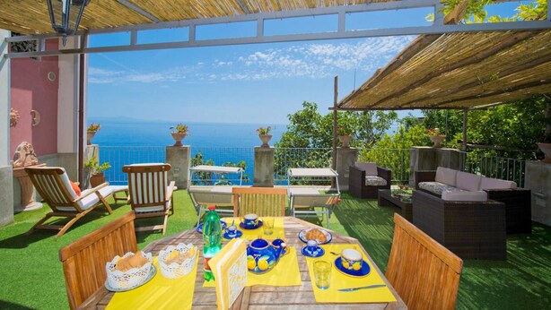 Un área de comedor al aire libre con vistas al mar, amueblada con sillas y mesas de madera, y una estructura de techo de paja.