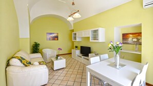 Una sala de estar con sofá, televisión, mesa de comedor y sillas. Se observan también armarios y una planta.