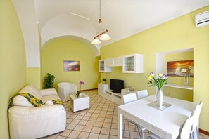 A living room with a sofa, TV, dining table, and chairs. There are cabinets and a plant. - Palazzo Rocco Villa Red Moon in Love – Sea View Terrace on the Amalfi Coast (Praiano)