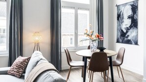 Ein modernes Wohnzimmer mit einer Couch, einem Esstisch mit Stühlen, einer Stehlampe und einem großen Fenster mit Vorhängen.