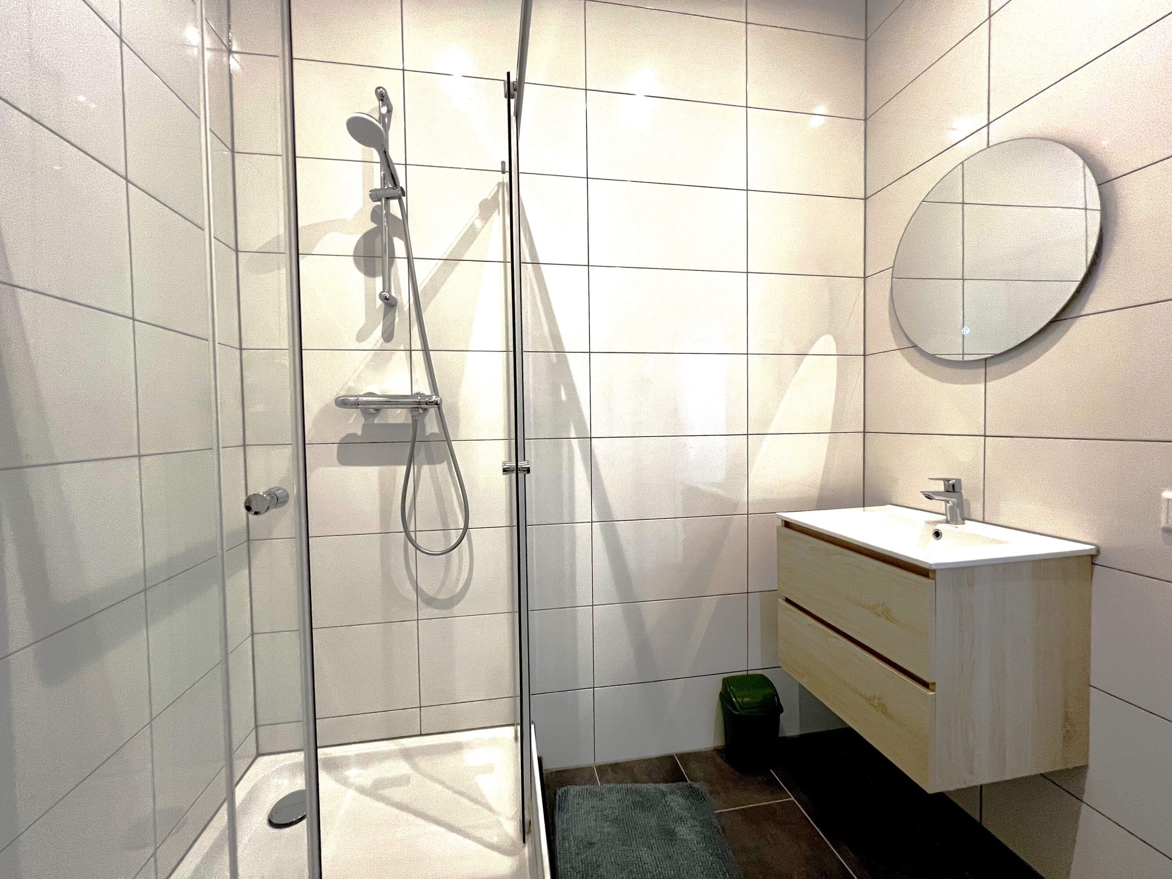 A modern bathroom with a glass-enclosed shower, a round mirror, and a wooden vanity.