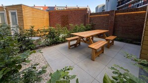 Eine Terrasse mit Holztischen und -bänken, umgeben von einer Backsteinmauer und Grünpflanzen.