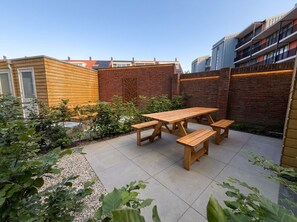 A patio with wooden tables and benches, surrounded by brick walls and greenery.