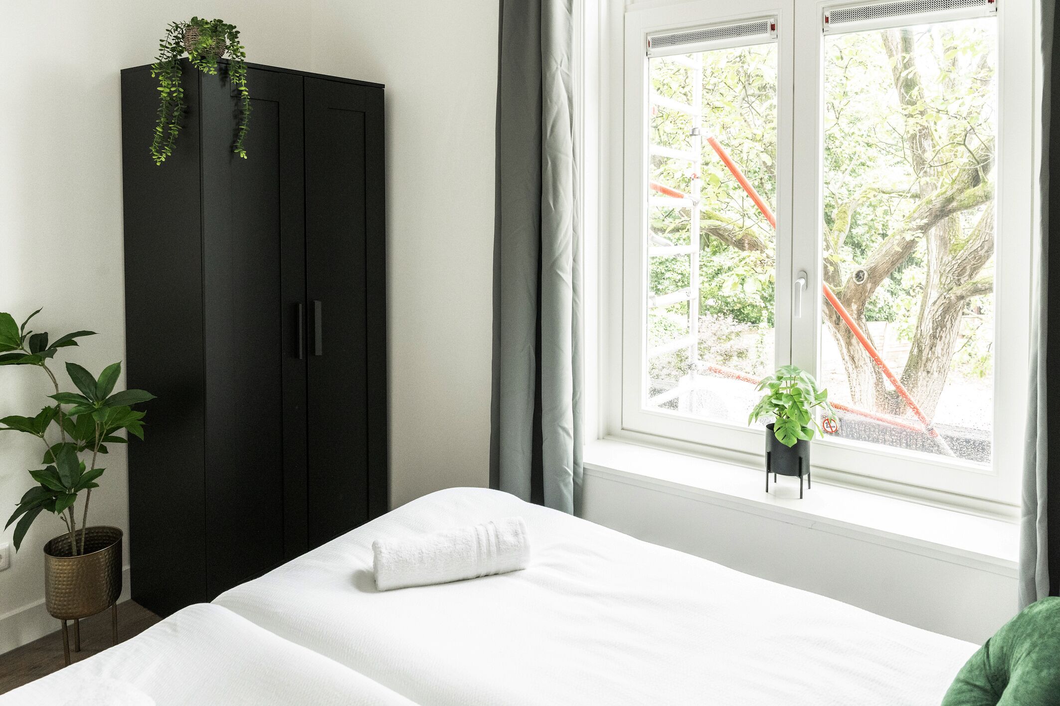 Un dormitorio con cama, un armario oscuro, una ventana con vista a árboles y una planta en maceta en el alféizar.