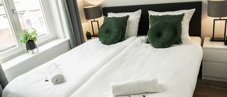 A double bed with white bedding and green pillows, a bedside table with a lamp, and a window with a view of buildings.