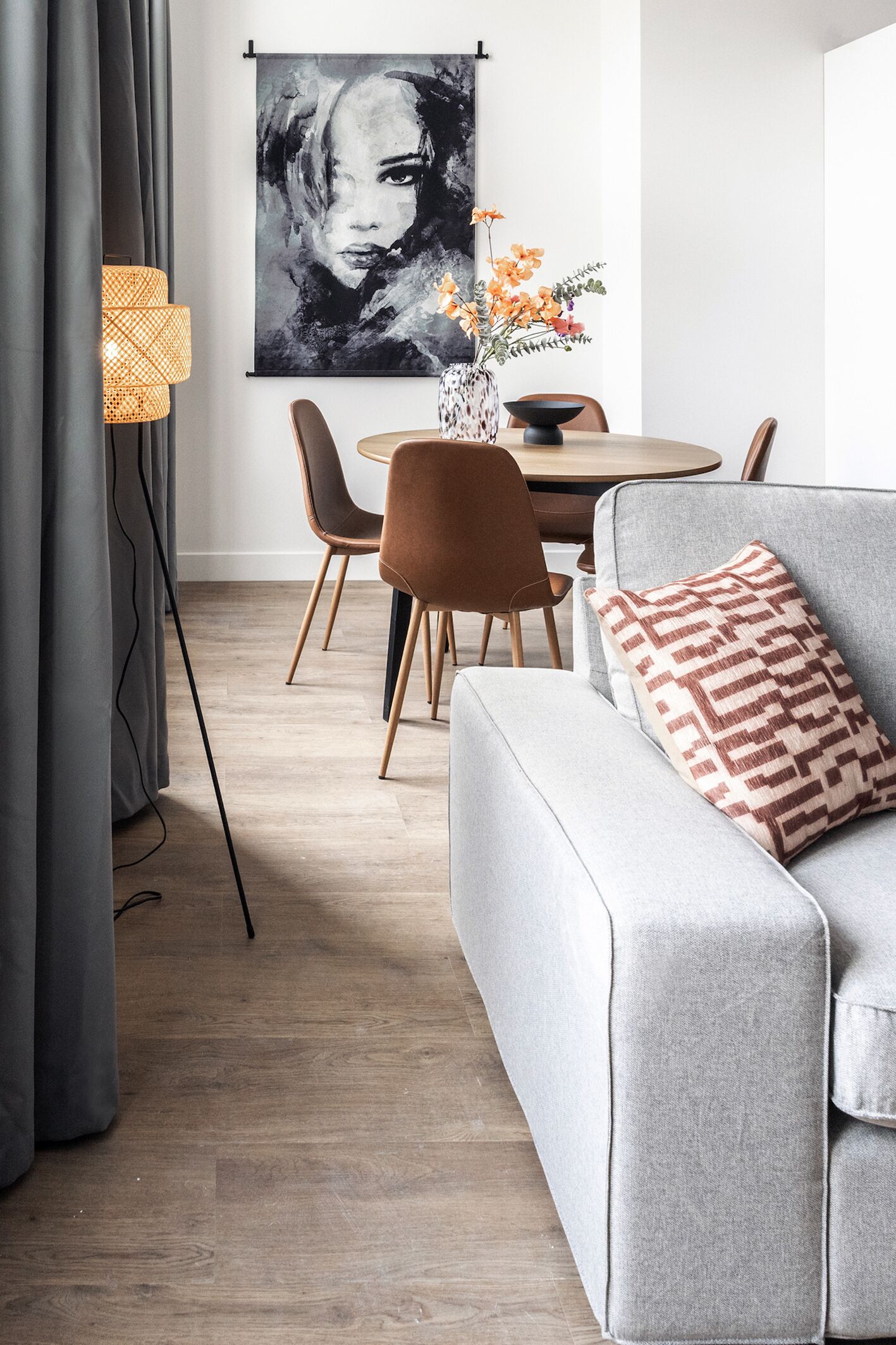 A modern living room with a grey sofa, a patterned throw pillow, a dining table with chairs, and a wall-mounted abstract painting.