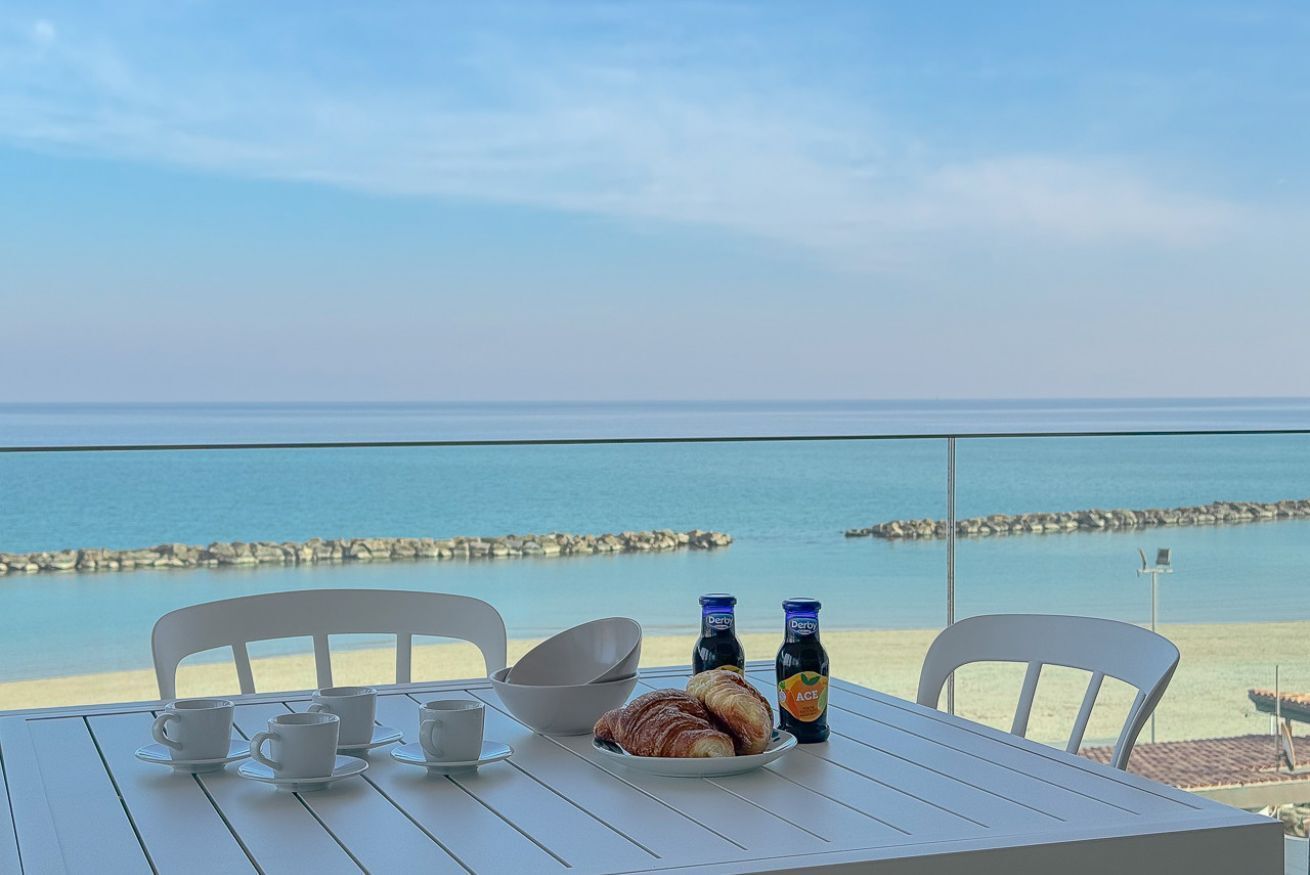 Ett bord med en tallrik full av croissanter, två ölflaskor och två koppar på en balkong med utsikt över en strand.