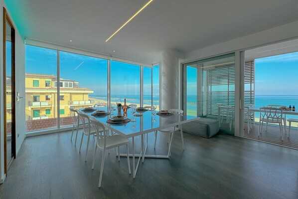 Zona pranzo moderna con un tavolo grande e sedie, ampie finestre vista mare e un balcone con posti a sedere all'aperto.