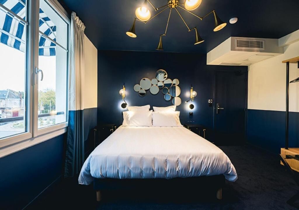 Une chambre à coucher avec un grand lit, un luminaire au plafond, un miroir fixé au mur et une fenêtre avec des rideaux.