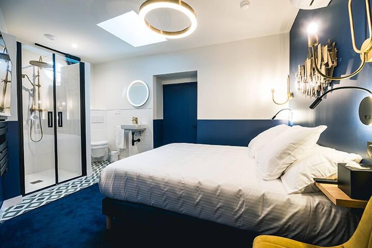 A modern hotel room with a large bed, a shower area, a small sink, and a round mirror.