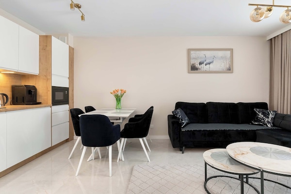 A bright living room with a compact dining area next to it. Light-colored walls and flooring contrast with a dark sofa and chairs. A minimalist design gives the space a clean and modern feel.