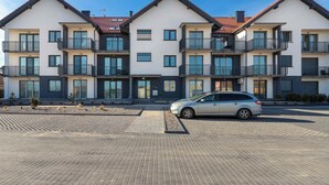 Un immeuble résidentiel à plusieurs étages doté de balcons, une voiture garée dans l’allée et une zone de stationnement dallée.