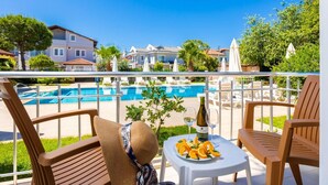 A balcony with a pool view, wooden chairs, a table with a bottle and glasses, and a plate of fruit.