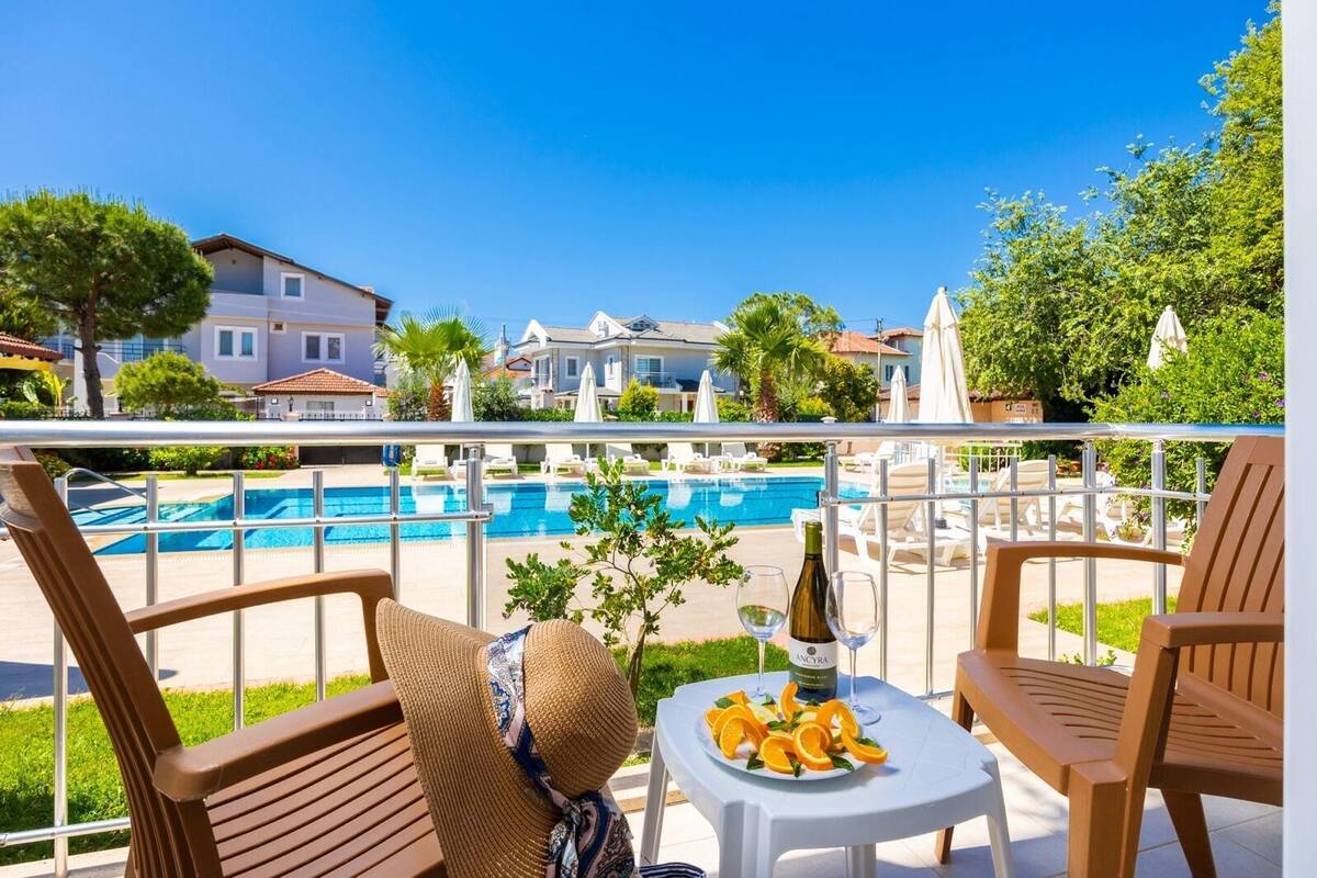Un balcon avec vue sur la piscine, des chaises en bois, une table avec une bouteille et des verres, et une assiette de fruits.