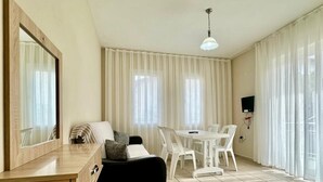 A room with a desk, a sofa, a dining table with chairs, a TV mounted on the wall, and large windows with curtains.