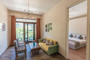 A living room with a sofa, a coffee table, a chair, a ceiling light fixture, and a view through a window. - Sojourn By the Lake - Boutique Hotel (Bhimtal)