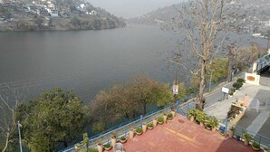 A river with a view of a hillside town, a railing with potted plants, and a clear sky. - Sojourn By the Lake - Boutique Hotel  (Bhimtal)