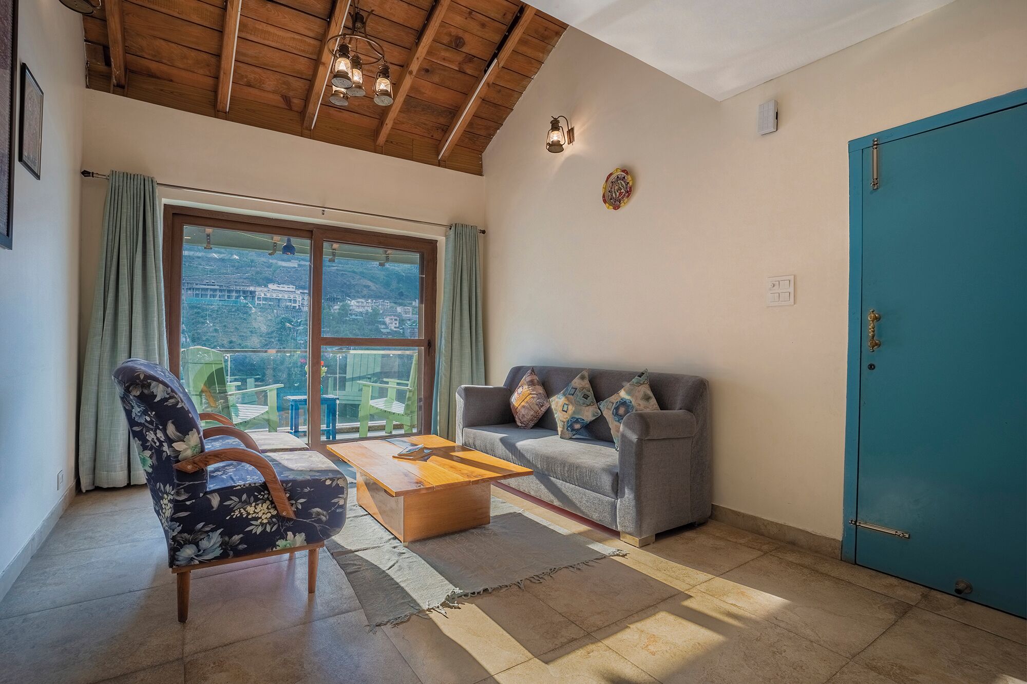 A room with a sofa, a chair, a wooden coffee table, a teal door, and a view through large windows.