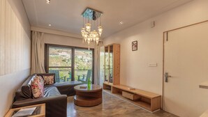 A modern living room with a sofa, a round wooden coffee table, a bench, and a view of a hillside. - Sojourn By the Lake - Boutique Hotel  (Bhimtal)