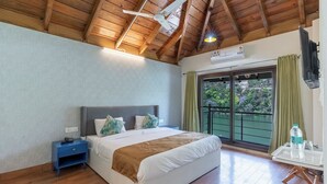 A bedroom with a bed, a nightstand, a ceiling fan, a window with curtains, and a view of greenery outside. - Sojourn By the Lake - Boutique Hotel  (Bhimtal)