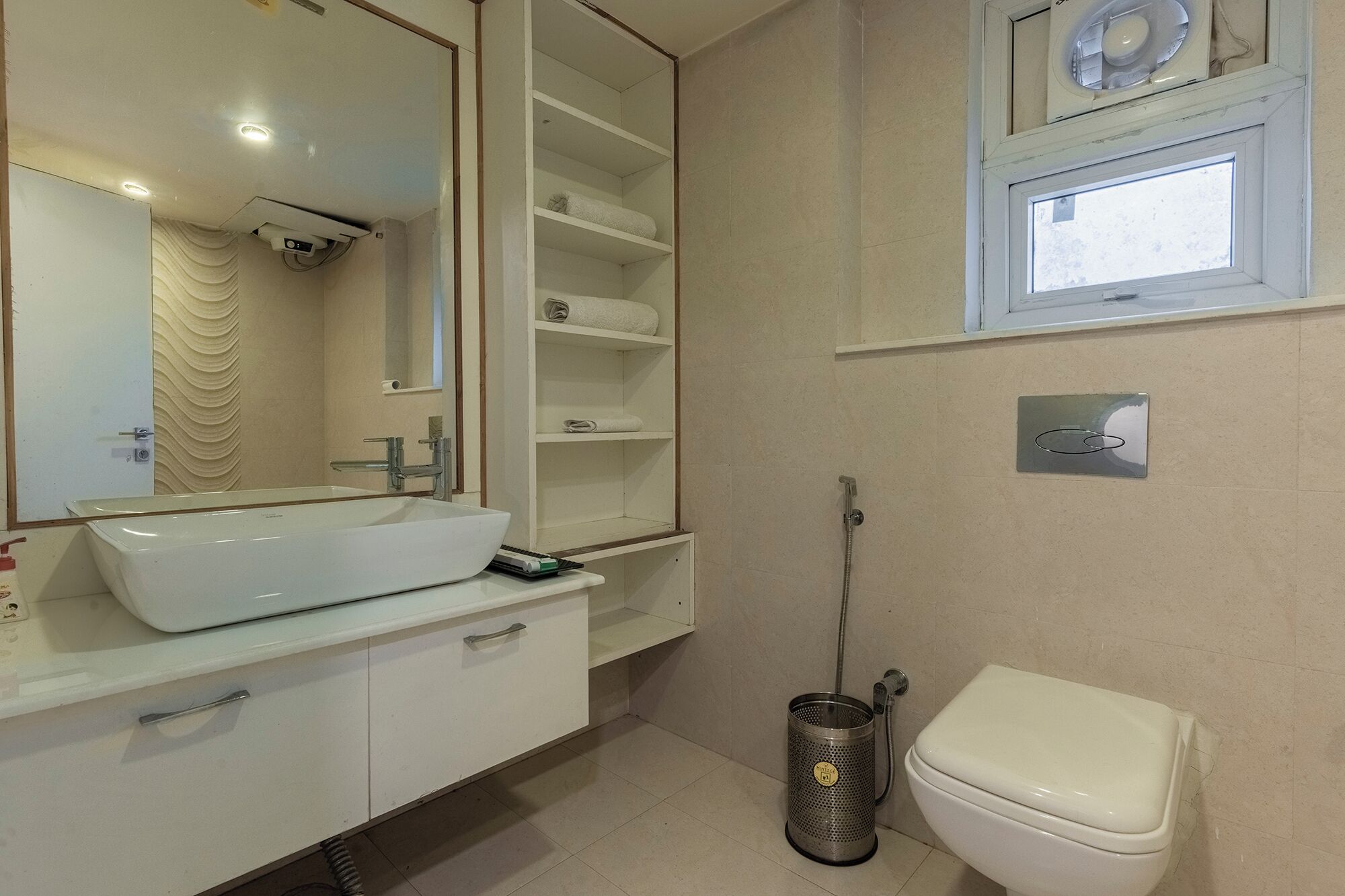 A bathroom with a white sink, mirror, and toilet. There are shelves above the sink and a window above the toilet.