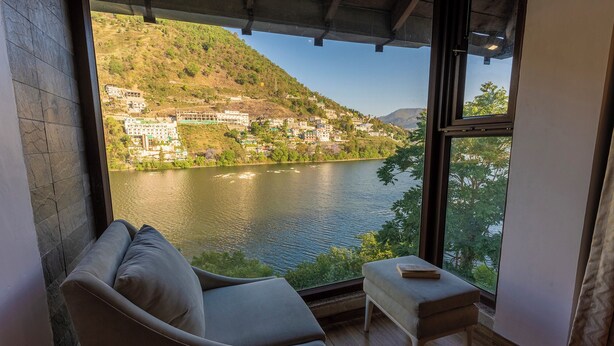 A room with a view of a lake and hills, a sofa, and a small table. - Sojourn By the Lake - Boutique Hotel  (Bhimtal)