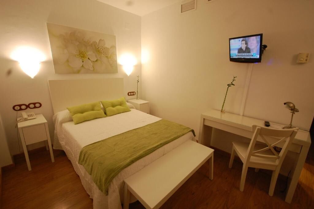 A bedroom with a bed, bedside table, TV, desk, chair, and a bench.