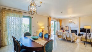 Uma área de refeições com mesa e cadeiras de madeira, um candelabro e vista pela janela com cortinas.