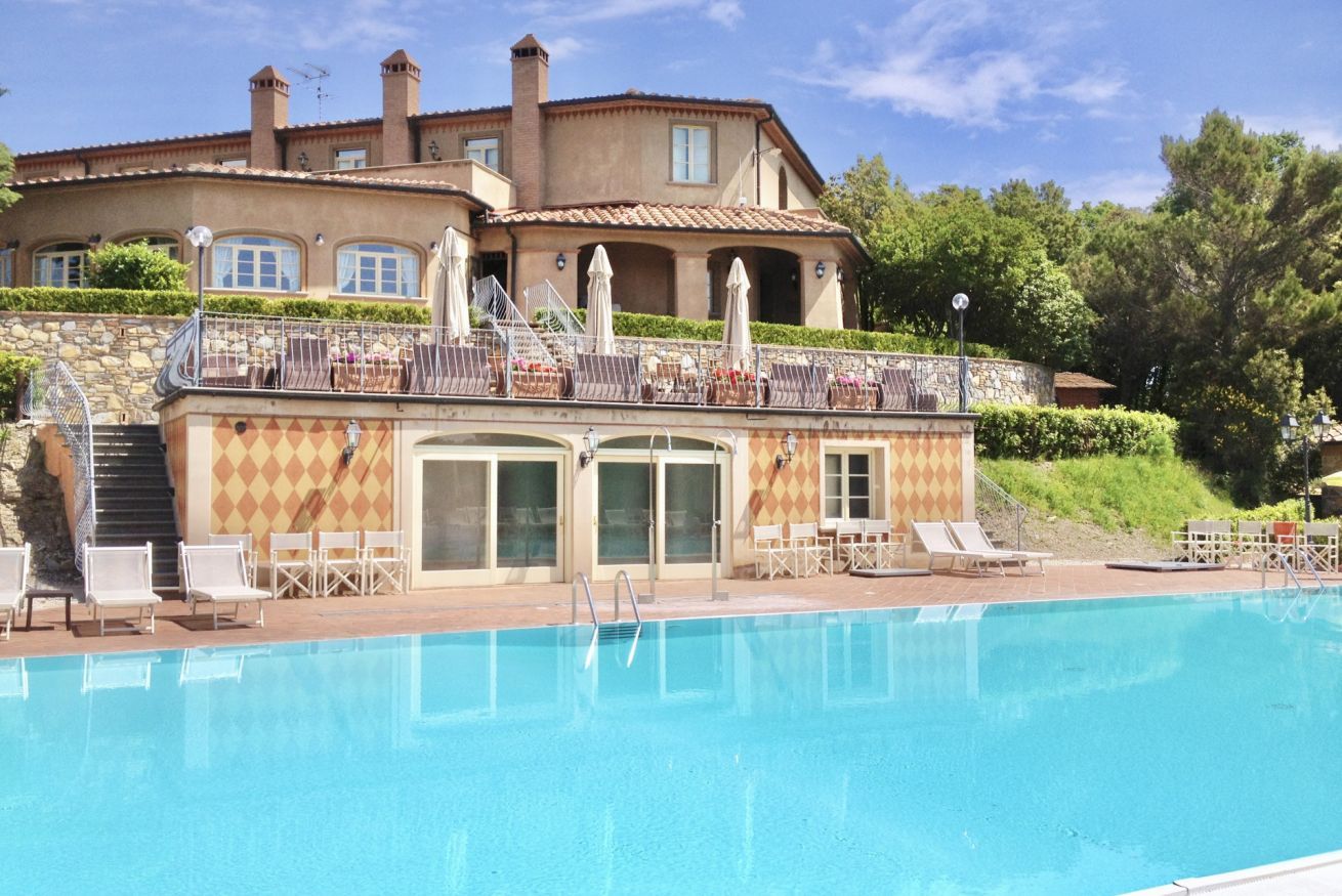 A pool area with lounge chairs, a building with a balcony, and a stone wall.