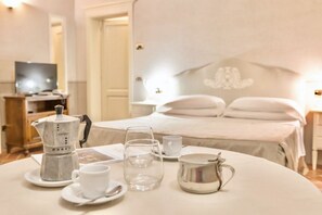 Ein ordentlich gedeckter Frühstückstisch mit Kaffeemaschine, Tassen und einer Kanne, vor einer Bettanlage und einem Fernseher in einem Hotelzimmer.