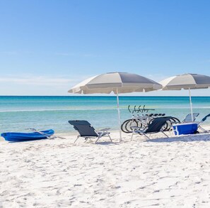 A beach with white sand, two beach chairs, a bicycle, and two white umbrellas.
