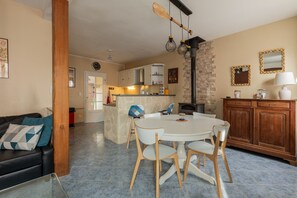 Un salon avec un canapé noir, une table basse en verre, une table à manger blanche avec des chaises, un coin cuisine avec des placards, un pilier en pierre et une cheminée.
