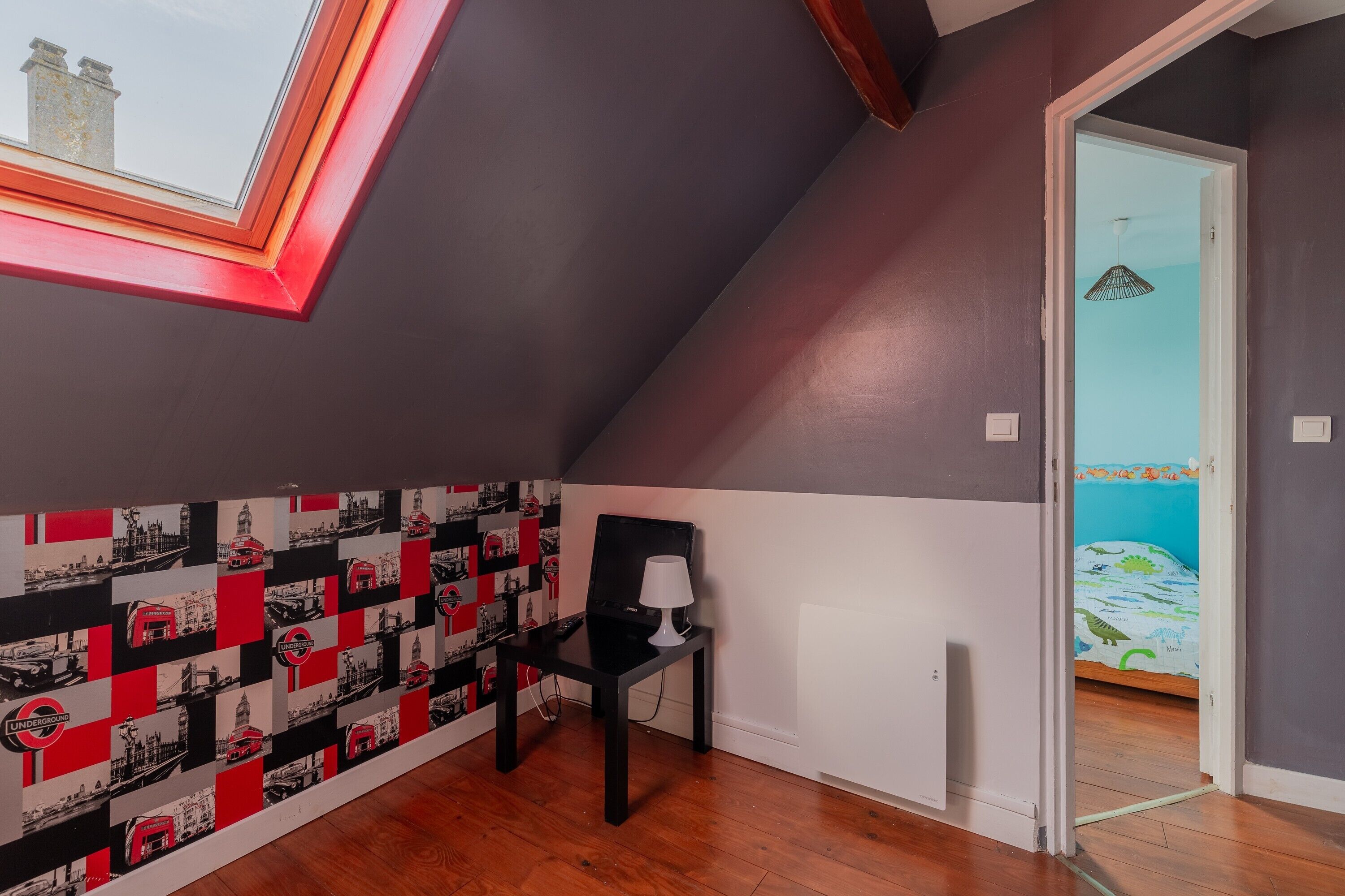 Una habitación con techo inclinado, un tragaluz, una silla negra, una lámpara blanca y una cama con edredón azul y verde.