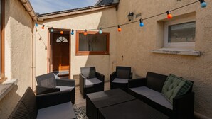 Une terrasse extérieure avec des meubles en osier, une petite fenêtre et des guirlandes lumineuses.
