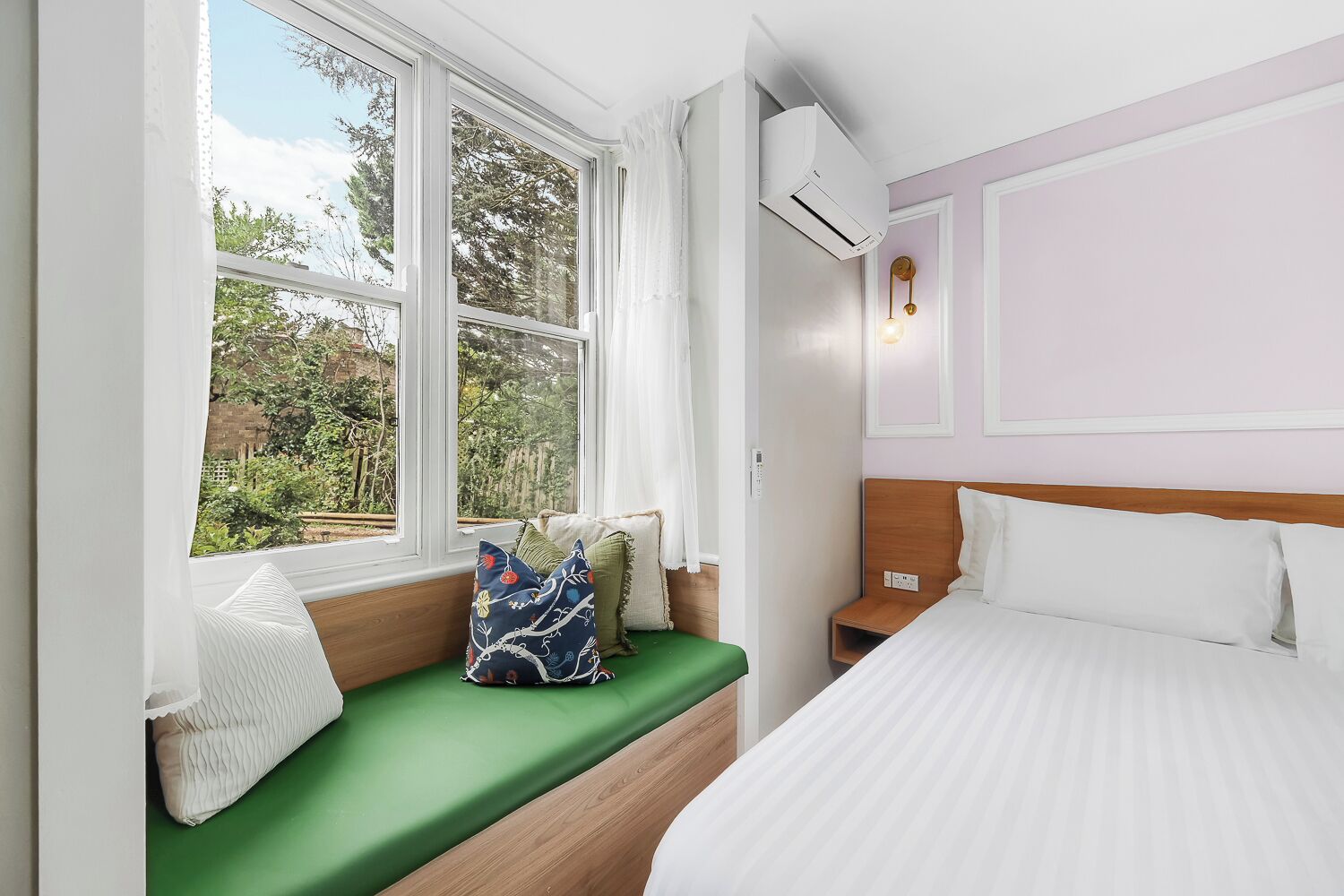 A bedroom with a bed, a green bench with pillows, a window with curtains, and an air conditioning unit.