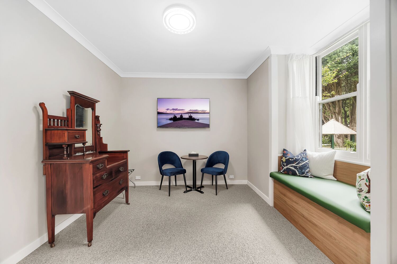 Um quarto com cômoda de madeira, duas cadeiras, uma mesa pequena, um banco verde com almofadas, um quadro e uma janela com vista para árvores.