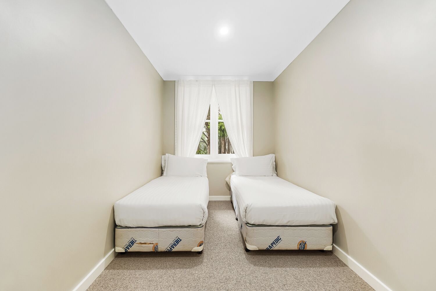 Quarto pequeno e estreito com duas camas de solteiro, uma janela com cortinas leves e vista para o verde lá fora.