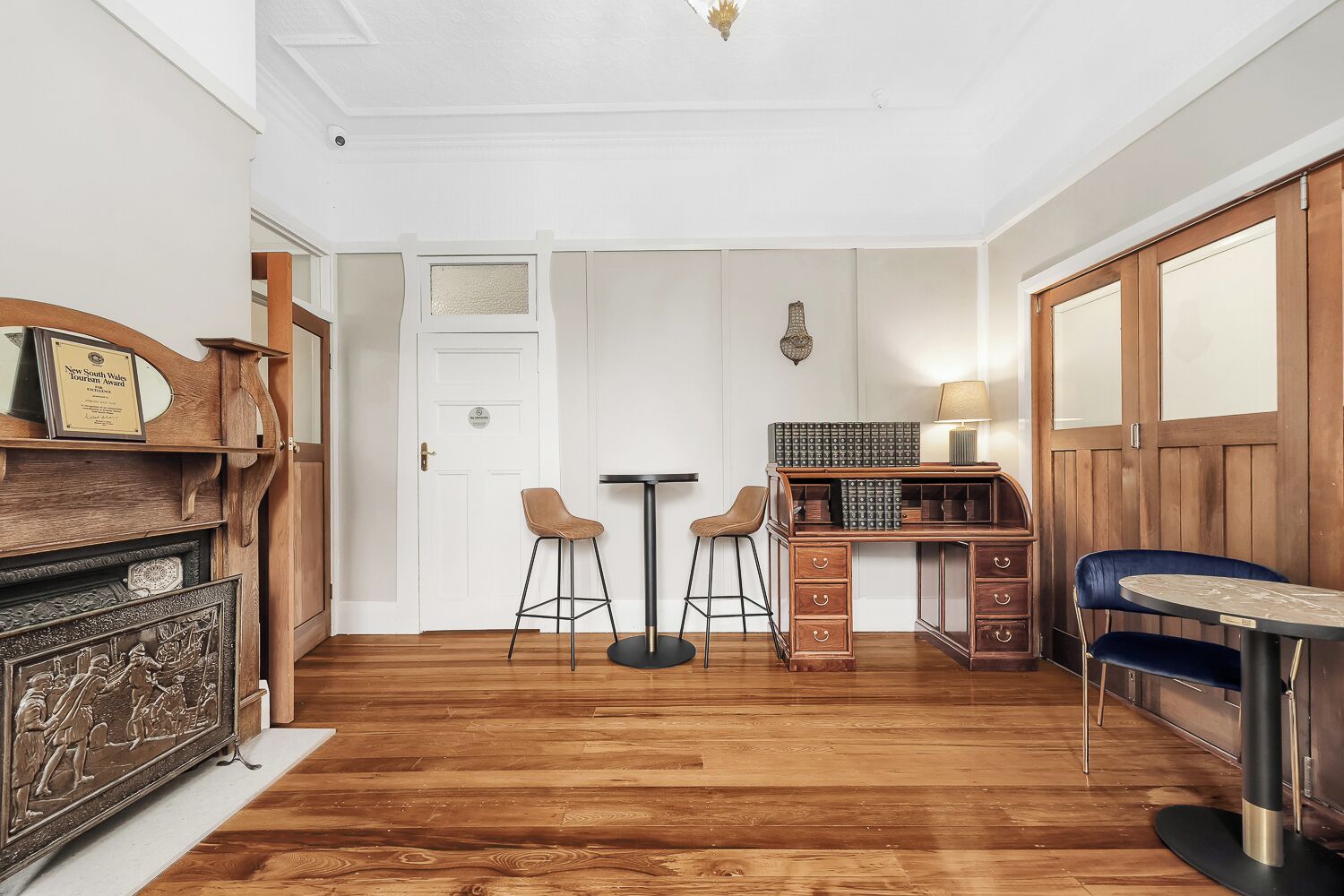 A room with wooden flooring, a desk with two chairs, a small table with a lamp, and a door with a plaque.