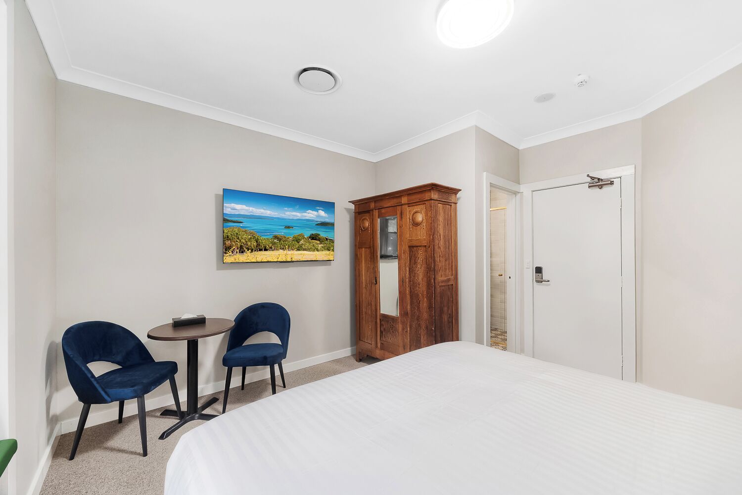A hotel room with a bed, two chairs, a small table, a wooden wardrobe, a painting, and a door.